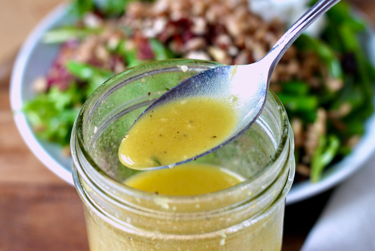 A metal spoon holding up some lemon thyme vinaigrette dressing above a glass jarful. Out of focus in the background is a small plate of salad.