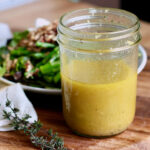 A glass jar filled with lemon thyme vinaigrette. Out of focus in the background is a plate of salad, a whole lemon, and a few sprigs of thyme.