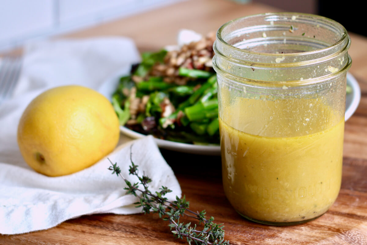 A glass jar filled with lemon thyme vinaigrette. Out of focus in the background is a plate of salad, a whole lemon, and a few sprigs of thyme.