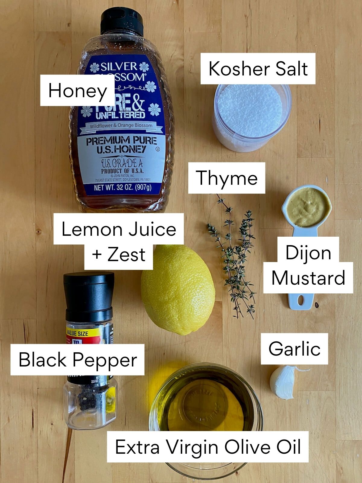 The ingredients to make lemon thyme vinaigrette dressing. Each ingredient is labeled with text. They include honey, kosher salt, thyme, lemon juice, lemon zest, dijon mustard, black pepper, garlic, and extra virgin olive oil.