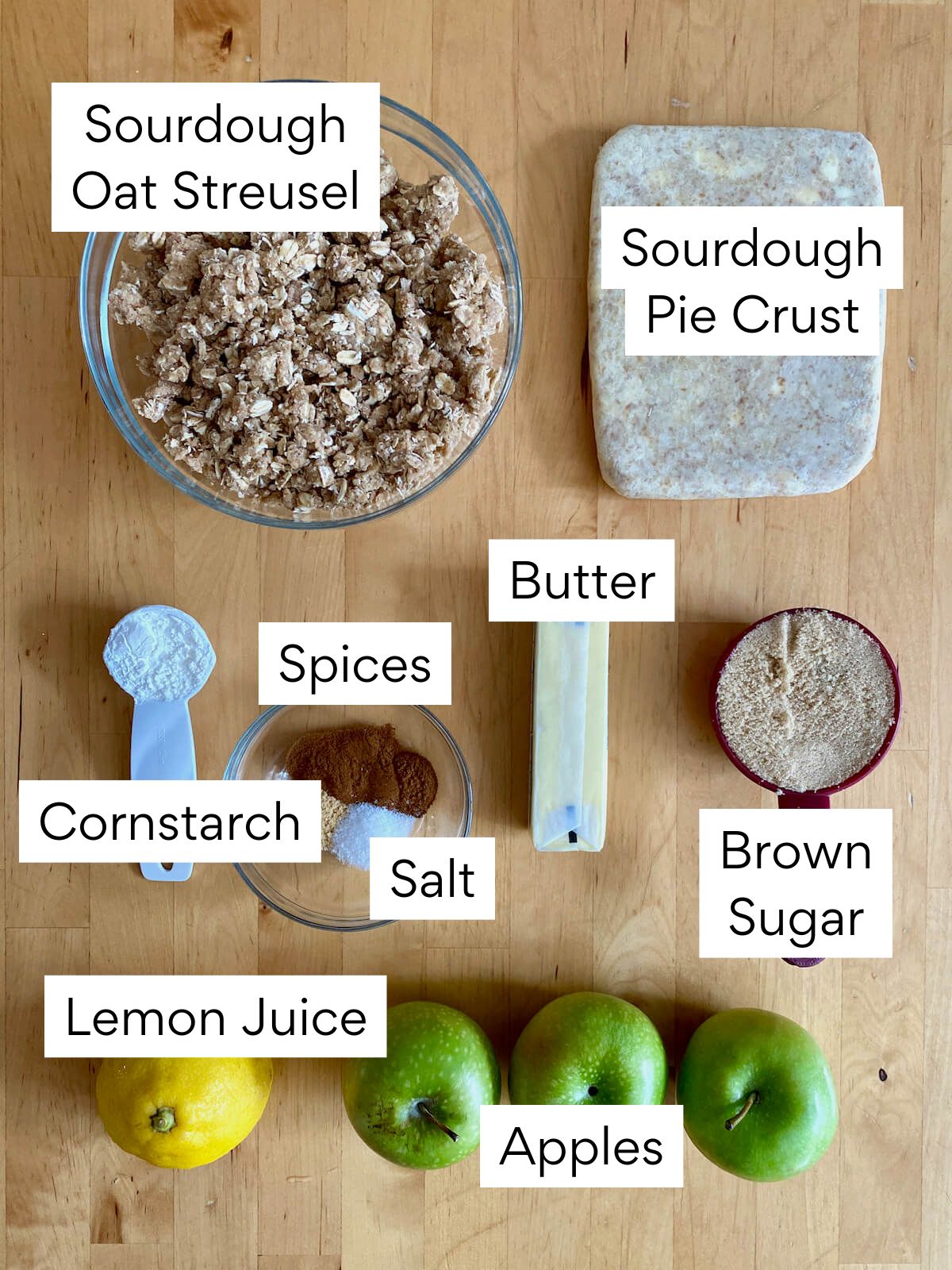The ingredients to make sourdough apple pie. Each ingredient is labeled with text. They include sourdough oat streusel, sourdough pie crust, butter, spices, salt, cornstarch, brown sugar, lemon juice, and apples.