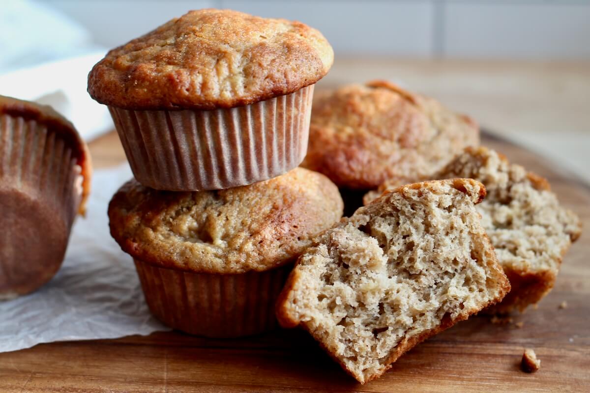 Several sourdough banana muffins stacked together. One of the muffins has been split in half to show the texture of the crumb.