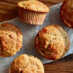 Five sourdough banana muffins on a piece of wrinkled parchment paper on top of a wooden cutting board.