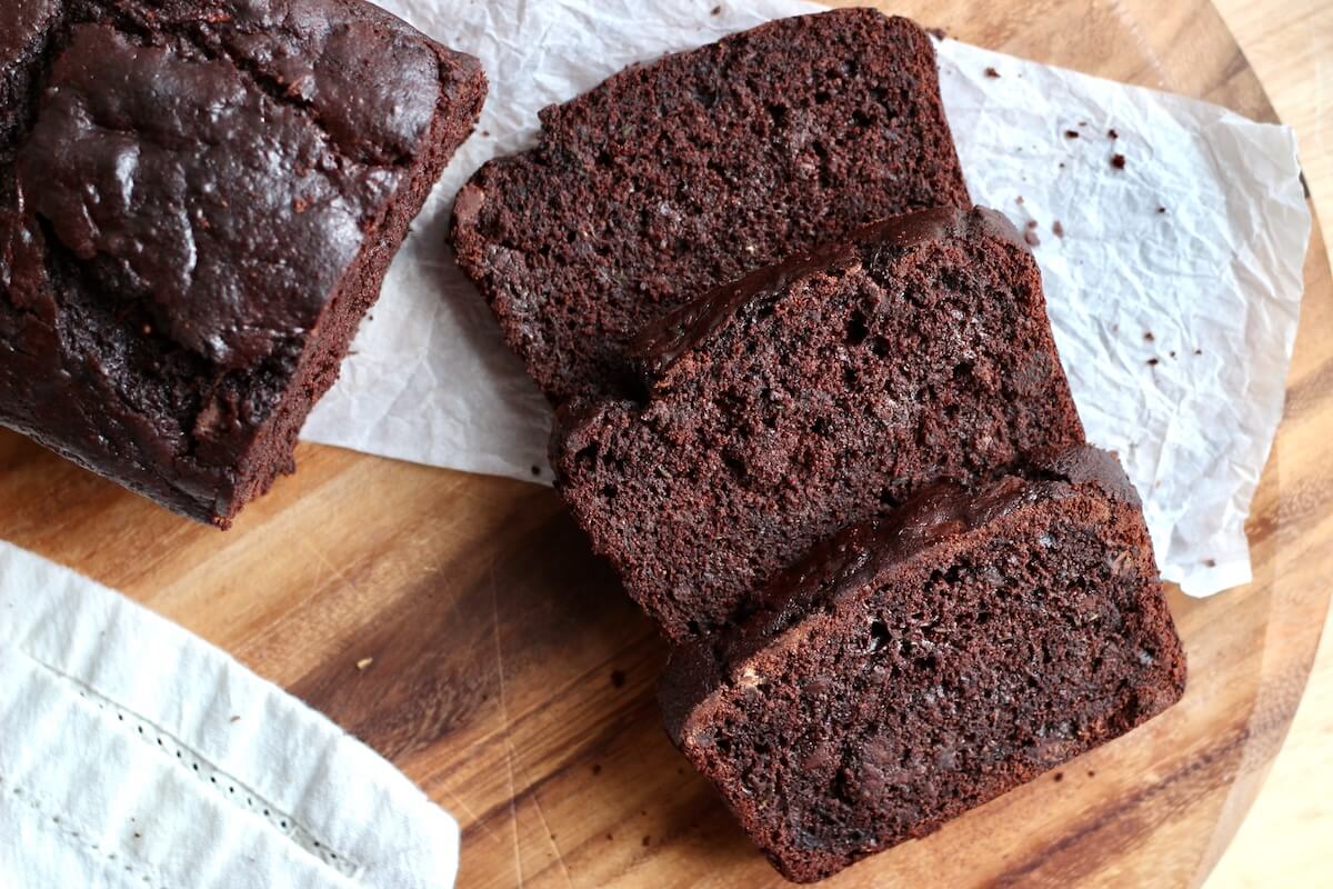 Three slices of sourdough chocolate zucchini bread laying on a wooden cutting board next to the rest of the loaf.