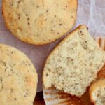 Sourdough lemon poppyseed muffins on a piece of wrinkled parchment paper. One of the muffins is split in half, laying on the paper liner.
