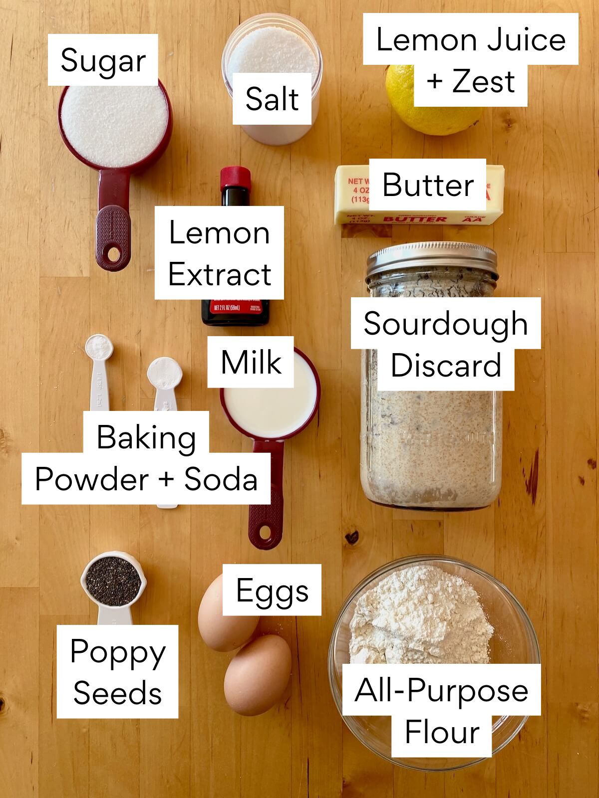 The ingredients to make sourdough lemon poppyseed muffins. Each ingredient is labeled with text. They include lemon juice, lemon zest, butter, sugar, salt, lemon extract, milk, sourdough discard, baking powder, baking soda, poppy seeds, eggs, and all-purpose flour.