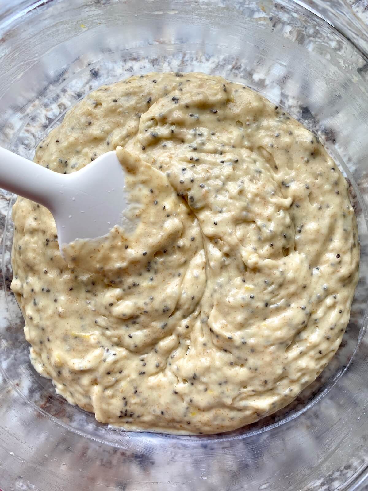 Sourdough lemon poppyseed muffin batter in a mixing bowl with a rubber spatula sticking out to the left.