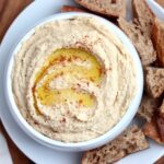 A bowl of hummus drizzled with olive oil and sprinkled with paprika surrounded by chunks of sourdough bread.