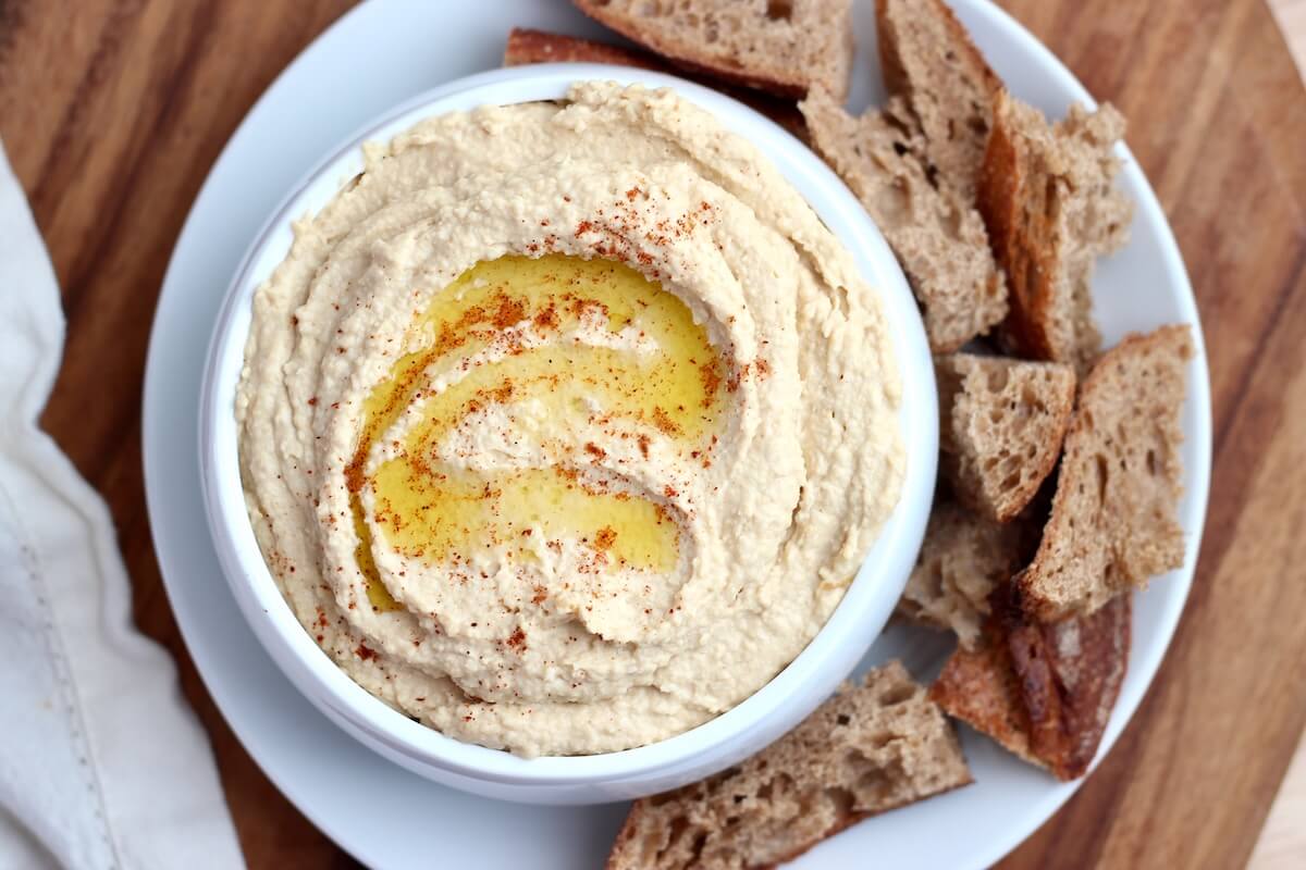 A bowl of hummus drizzled with olive oil and sprinkled with paprika surrounded by chunks of sourdough bread.