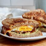 A bacon, egg, and cheese sourdough breakfast sandwich with avocado cut in half on a small white plate.