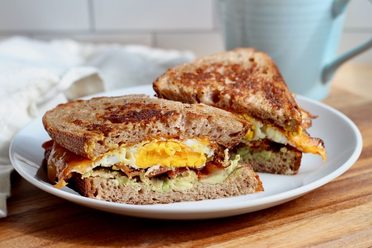 A bacon, egg, and cheese sourdough breakfast sandwich with avocado cut in half on a small white plate.