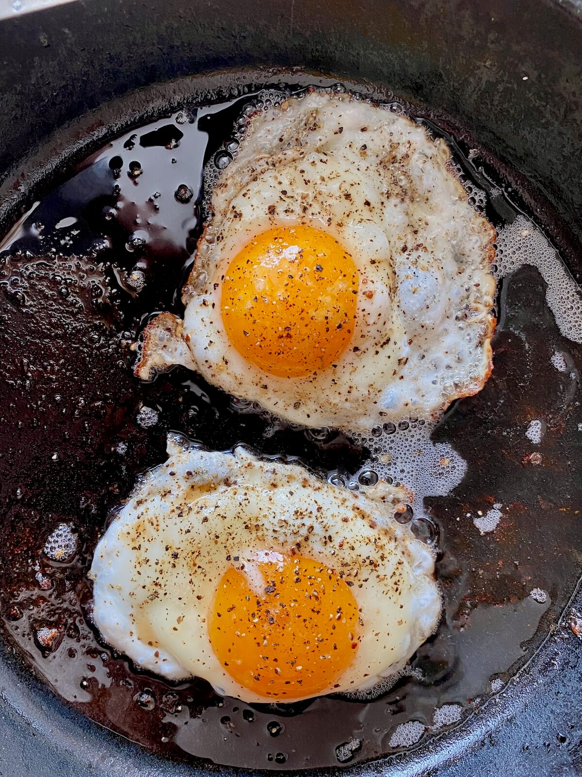 Two eggs seasoned with salt and pepper frying in bacon fat in a cast iron skillet.