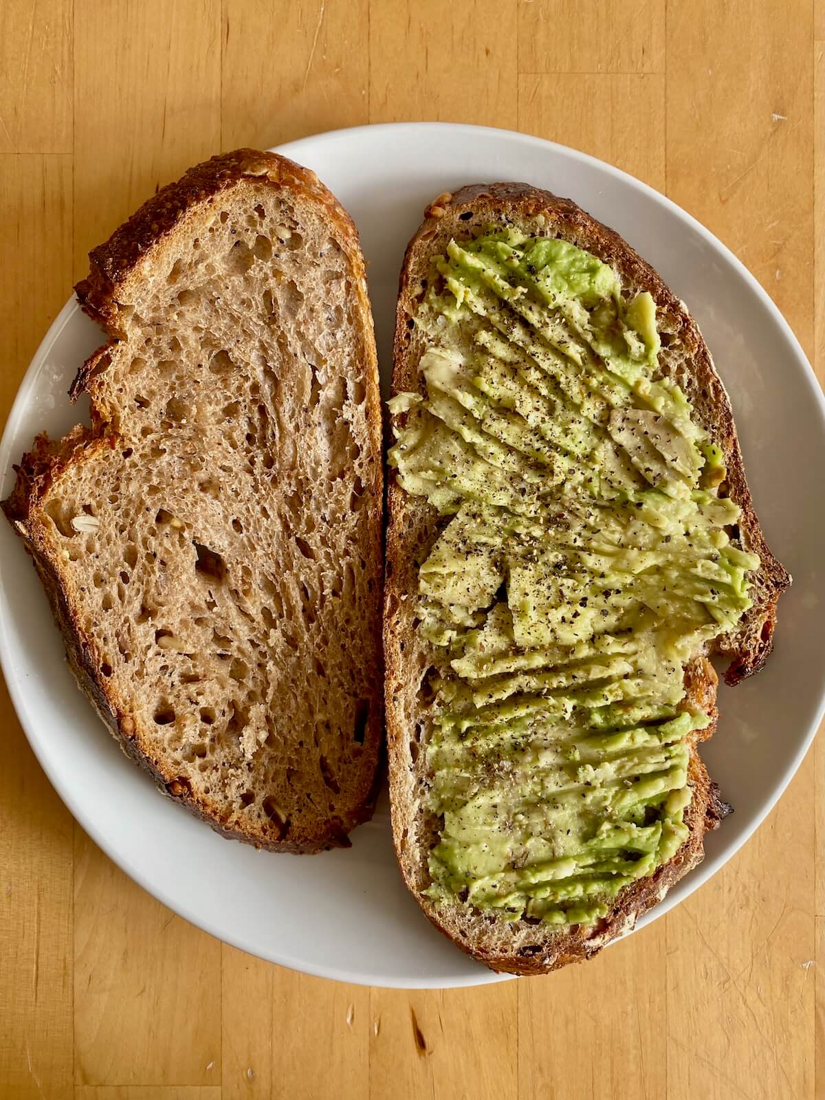 Two slices of toasted sourdough bread on a small white plate. The slice of the left is covered with mashed avocado that's been seasoned with salt and pepper.