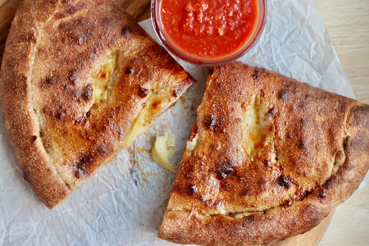 A pepperoni sourdough calzone split in two on a sheet of parchment paper next to a small bowl of marinara sauce.