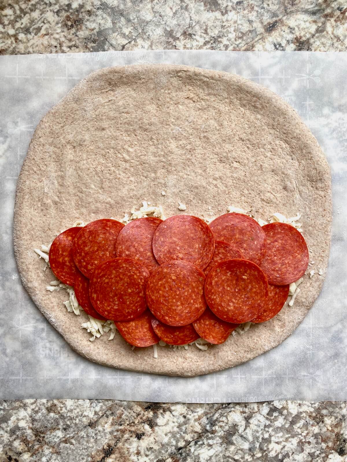 Pepperoni layered on top of cheese on half of a stretched out sourdough pizza dough.
