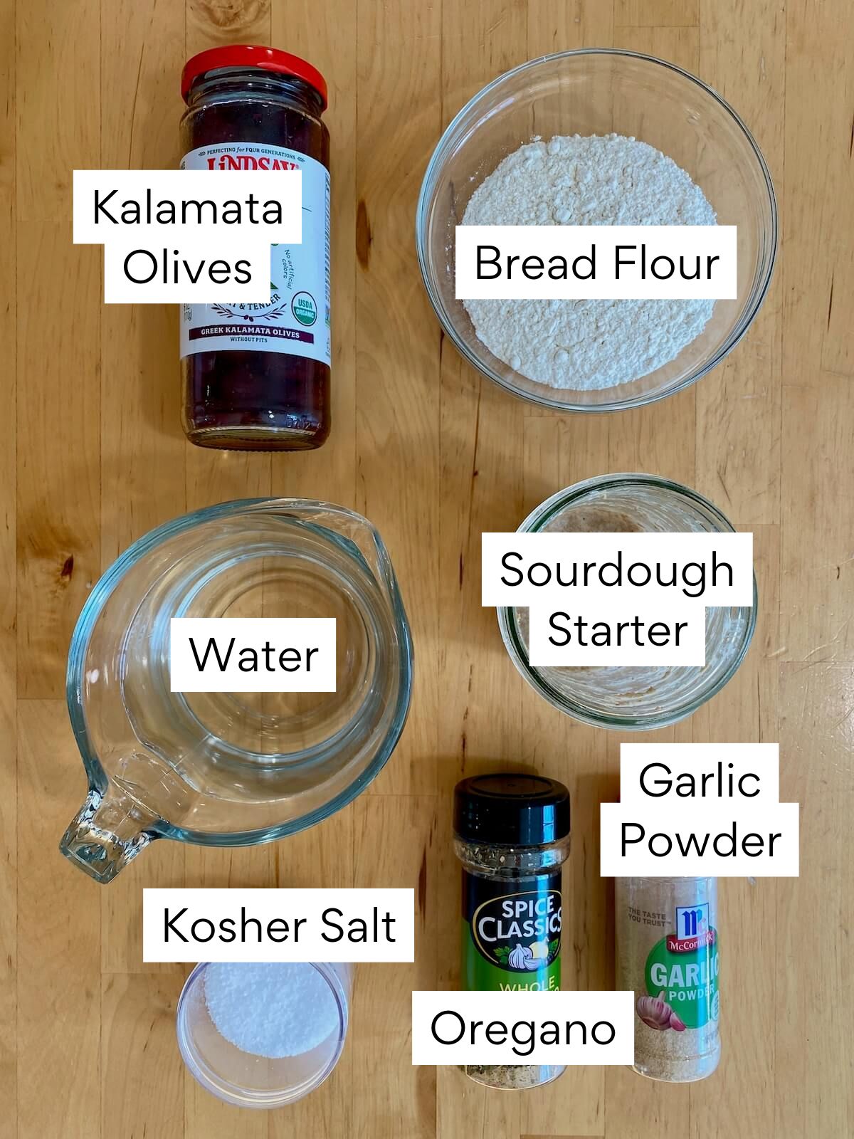 The ingredients to make sourdough olive bread. Each ingredient is labeled with text. They include kalamata olives, bread flour, water, sourdough starter, kosher salt, oregano, and garlic powder.