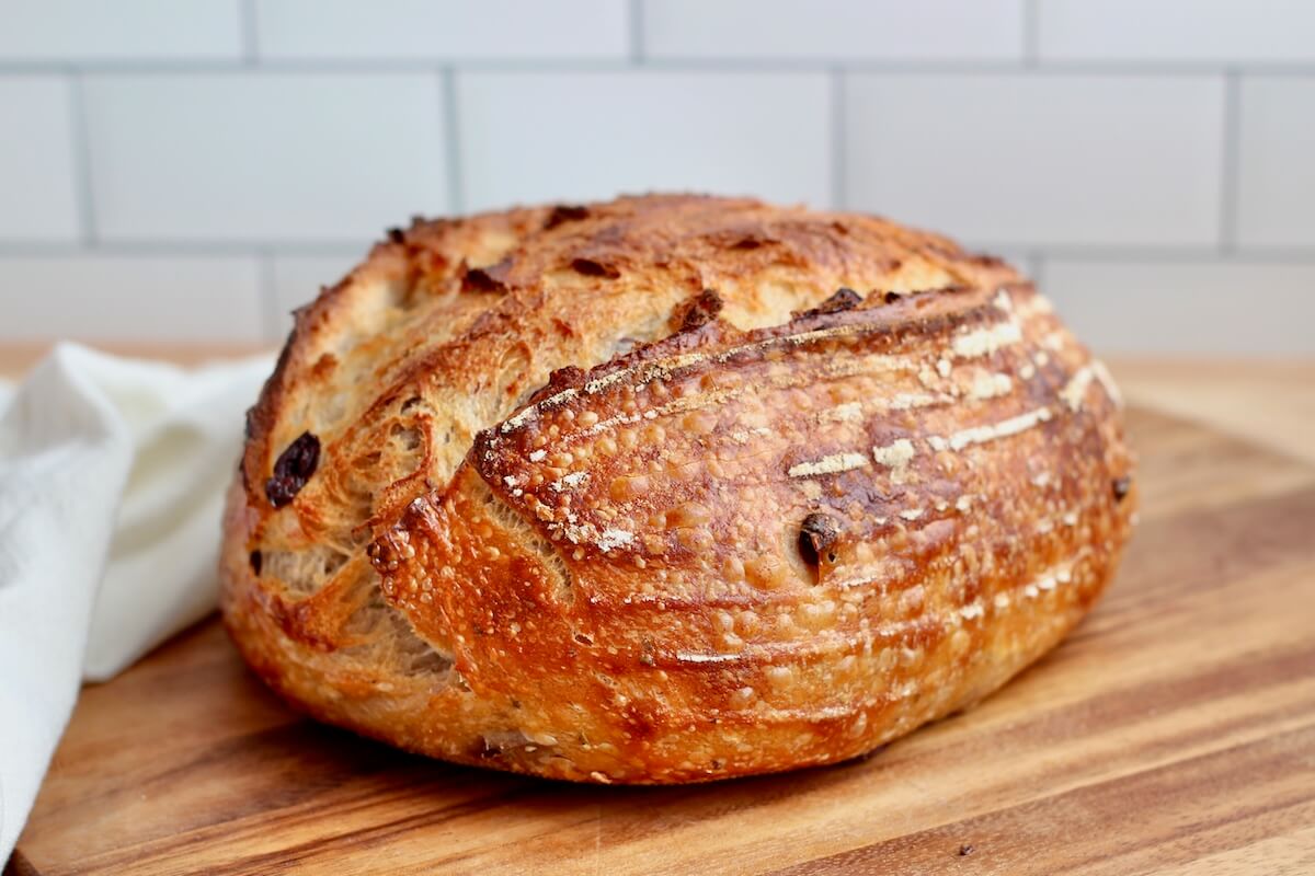 A loaf of olive sourdough bread on a wooden cutting board next to a white cloth napkin.