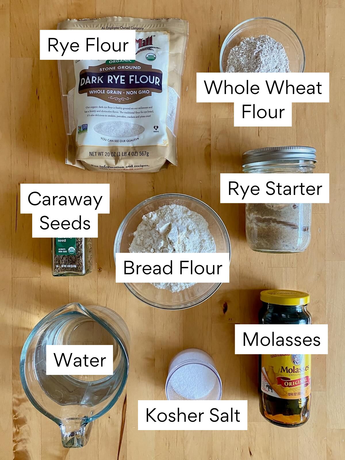 The ingredients to make sourdough rye bread. Each ingredient is labeled with text. They include rye flour, whole wheat flour, rye starter, bread flour, caraway seeds, water, kosher salt, and molasses.