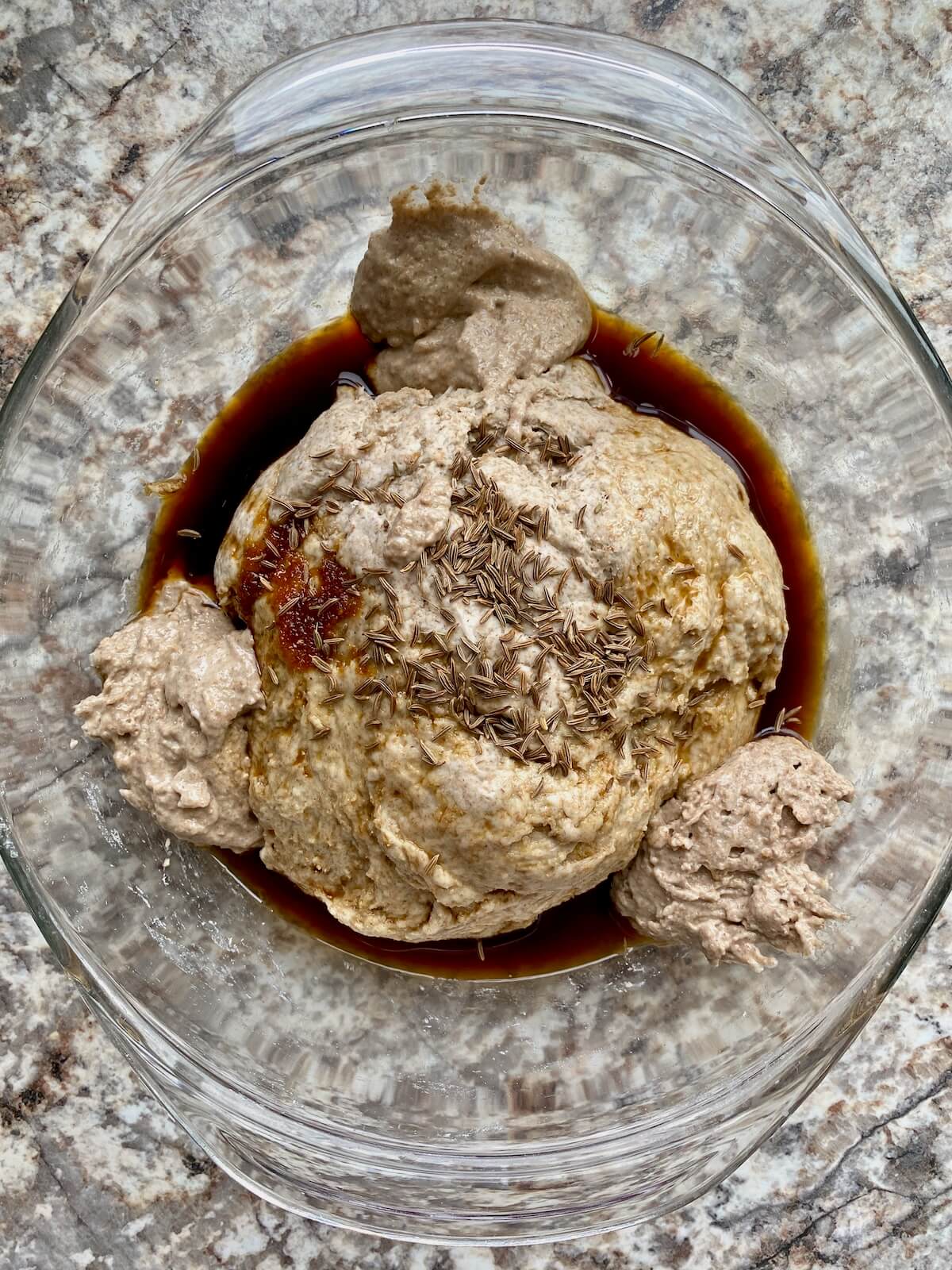 The starter, molasses, and caraway seeds added to the flour water mixture in a glass bowl.
