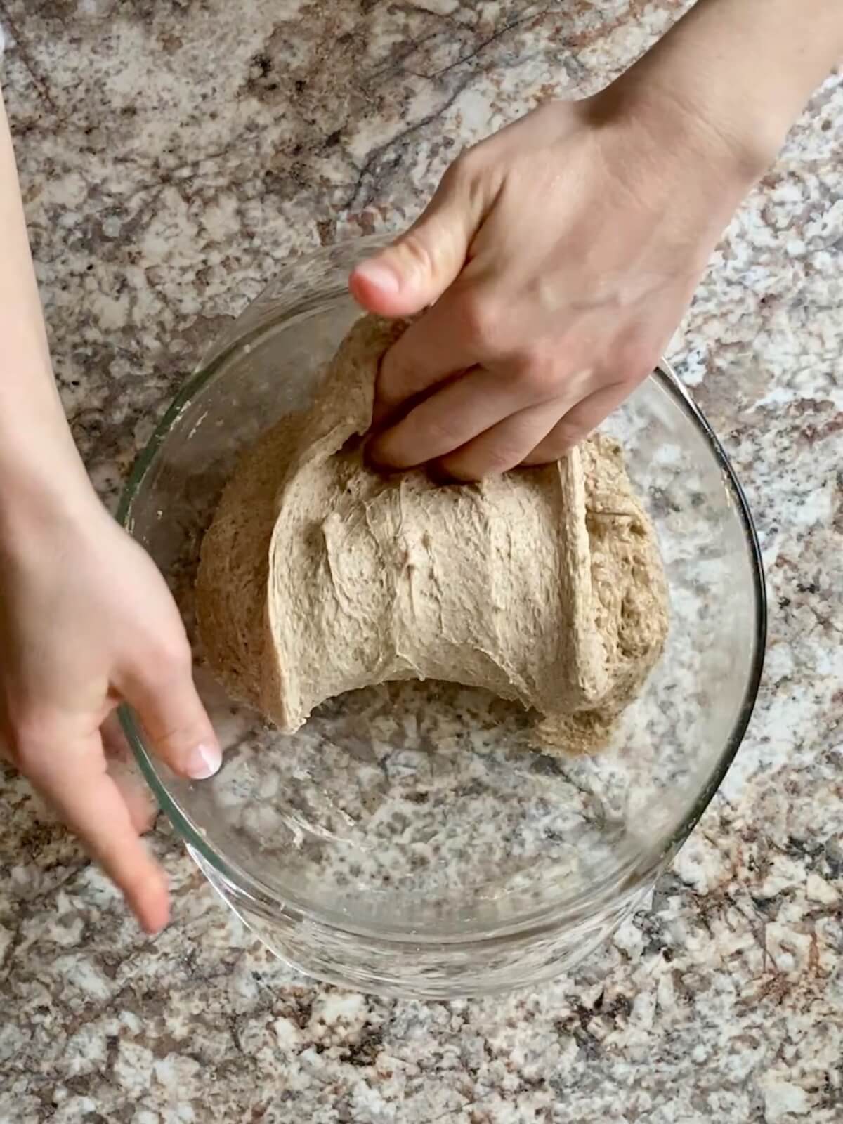 Two hands stretching and folding the sourdough rye bread dough in a glass bowl.