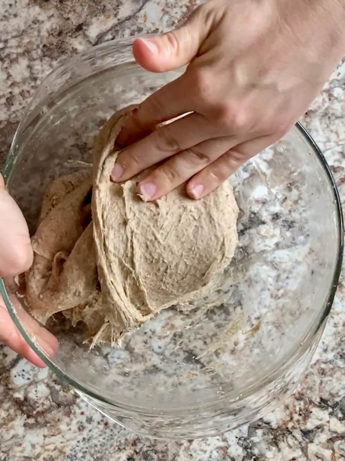 Two hands stretching and folding the sourdough rye bread dough in a glass bowl.