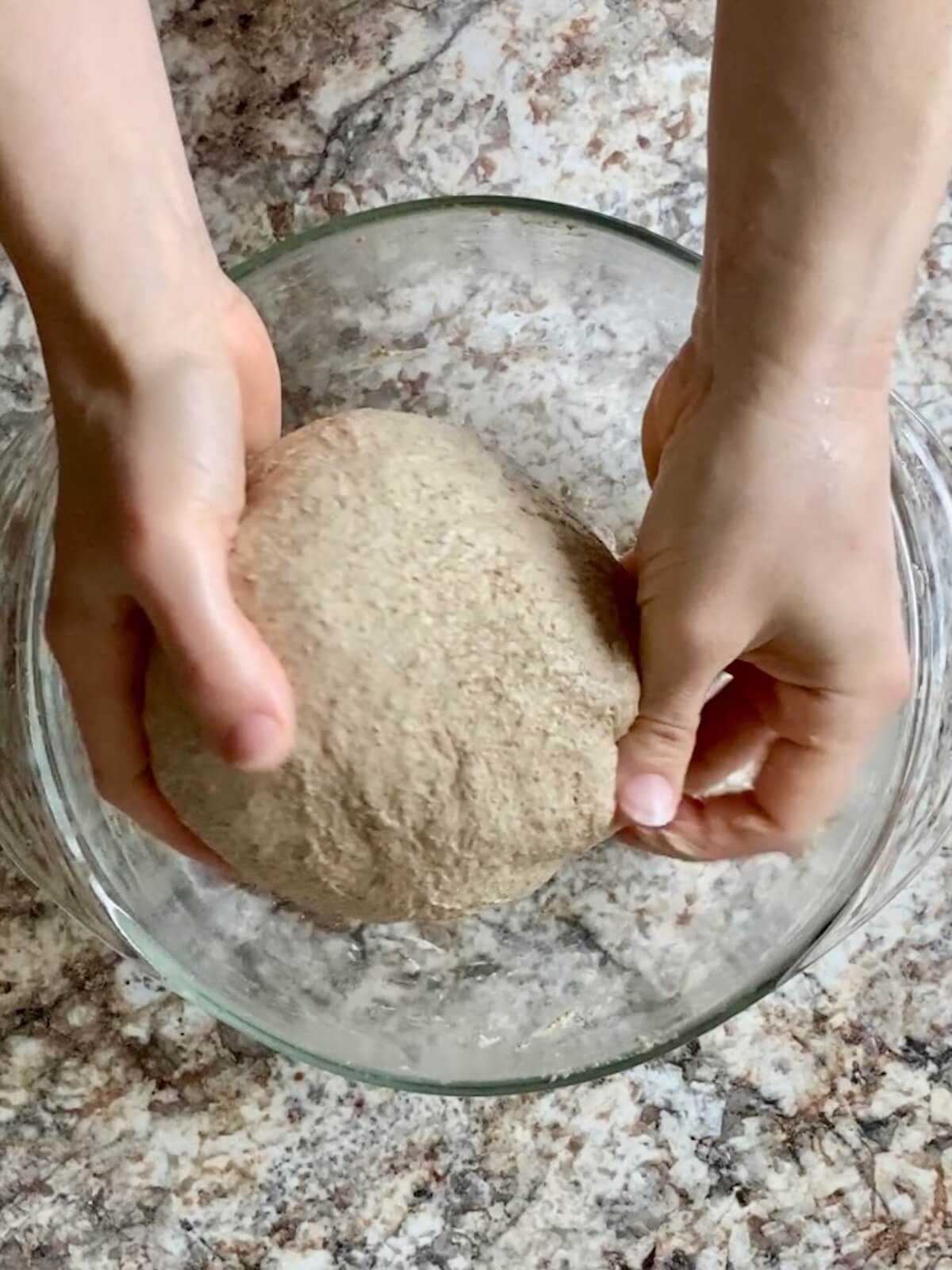 The rye sourdough bread dough being shaped into a ball by two hands above a glass bowl.