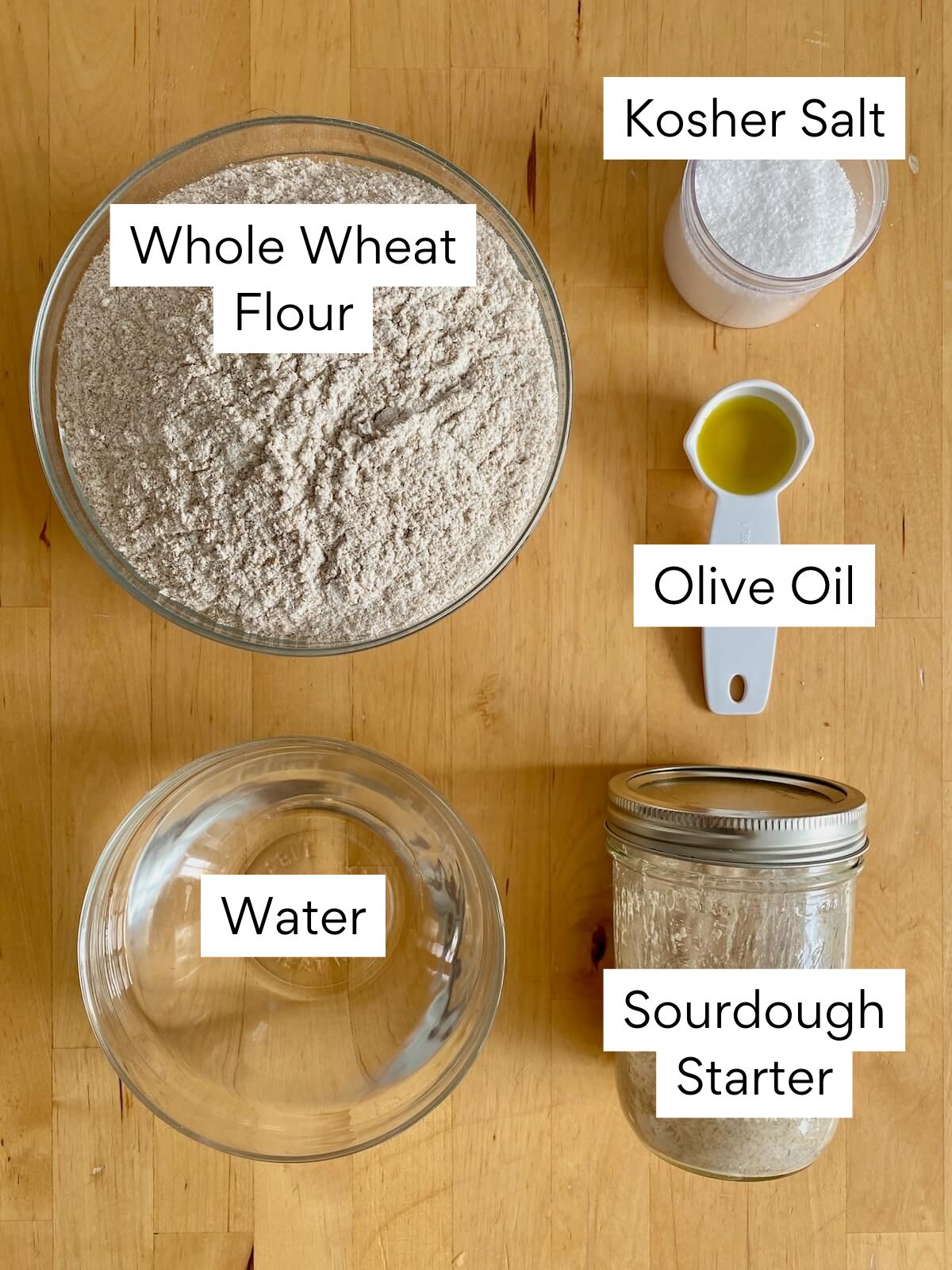 The ingredients to make whole wheat sourdough focaccia. Each ingredient is labeled with text. They include whole wheat flour, kosher salt, olive oil, water, and sourdough starter.