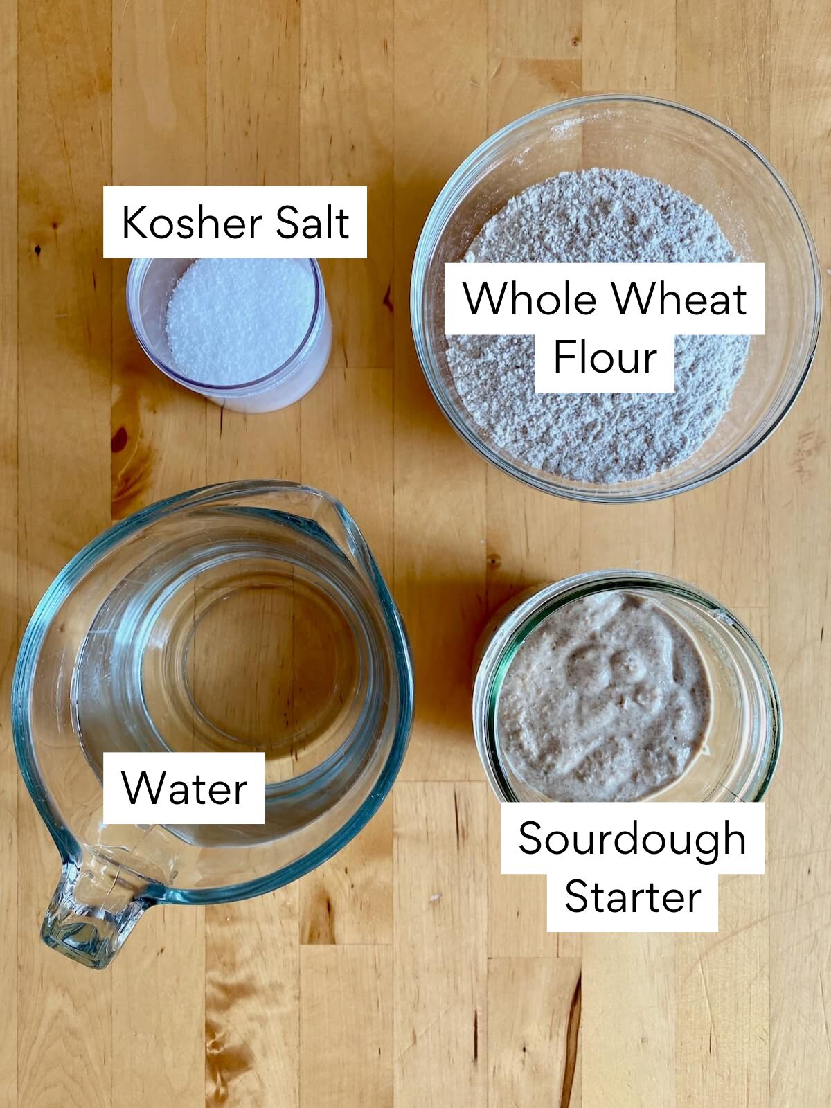 The ingredients to make sourdough whole wheat pizza dough. Each ingredient is labeled with text. They include whole wheat flour, kosher salt, water, and sourdough starter.
