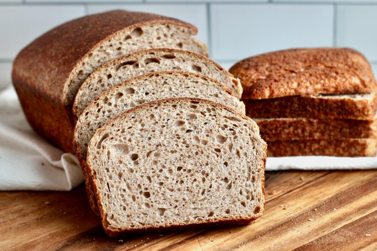 A loaf of whole wheat sourdough sandwich bread cut into slices on a wooden cutting board.