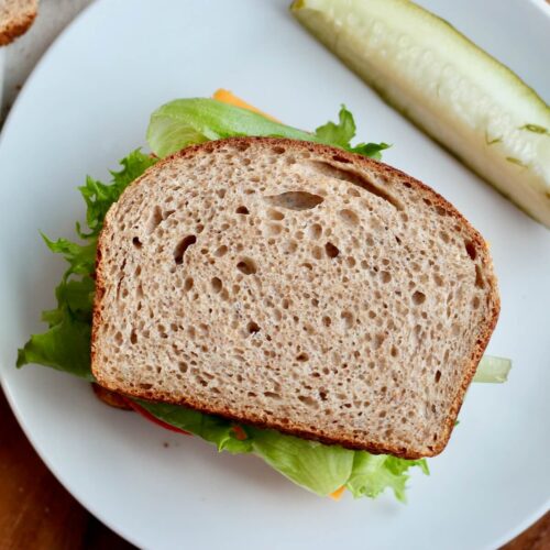 A sandwich made on whole wheat sourdough sandwich bread. The sandwich is on a white plate and their is a pickle spear next to it.