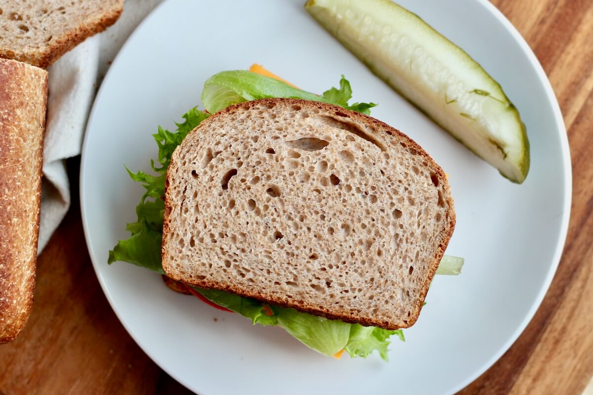 A sandwich made on whole wheat sourdough sandwich bread. The sandwich is on a white plate and their is a pickle spear next to it.