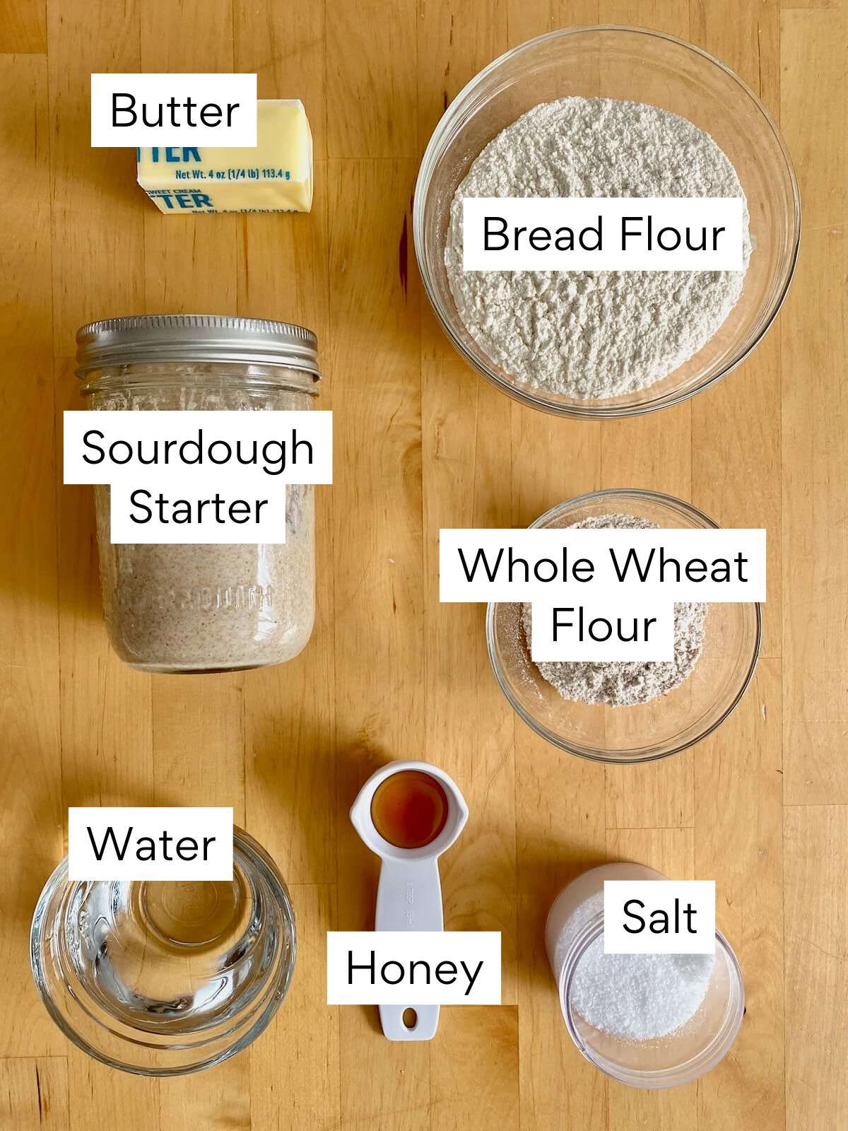 The ingredients to make whole wheat sourdough sandwich bread. Each ingredient is labeled with text. They include butter, bread flour, sourdough starter, whole wheat flour, water, honey, and salt.