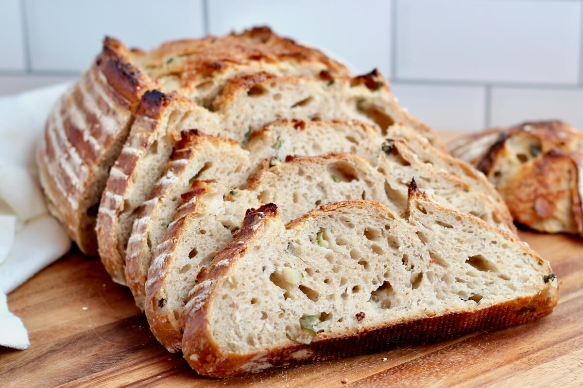 A loaf of dill pickle sourdough bread cut into slices on a wooden cutting board.