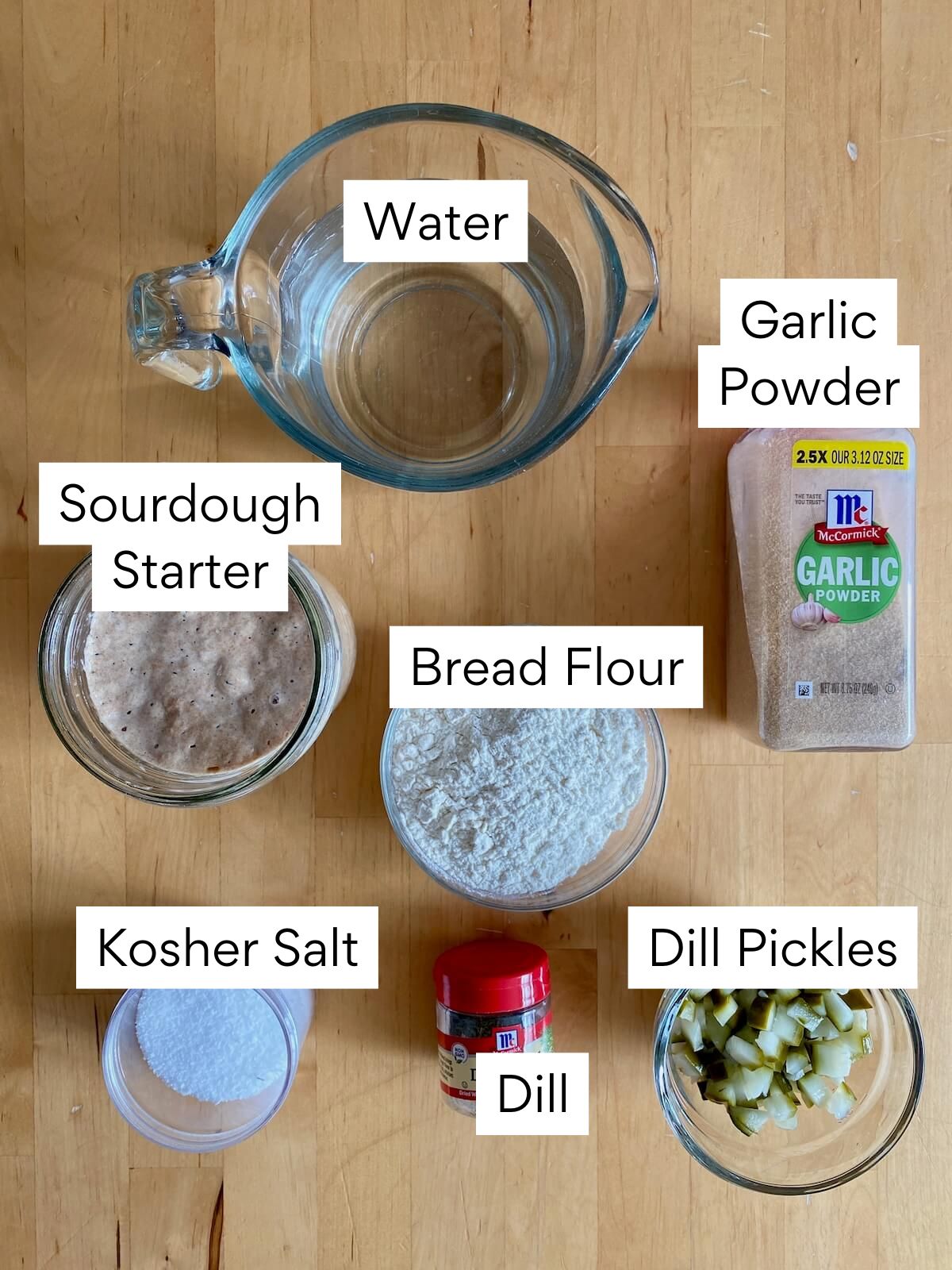 The ingredients to make pickle sourdough bread. Each ingredient is labeled with text. They include water, sourdough starter, bread flour, kosher salt, dill, garlic powder, and dill pickles.