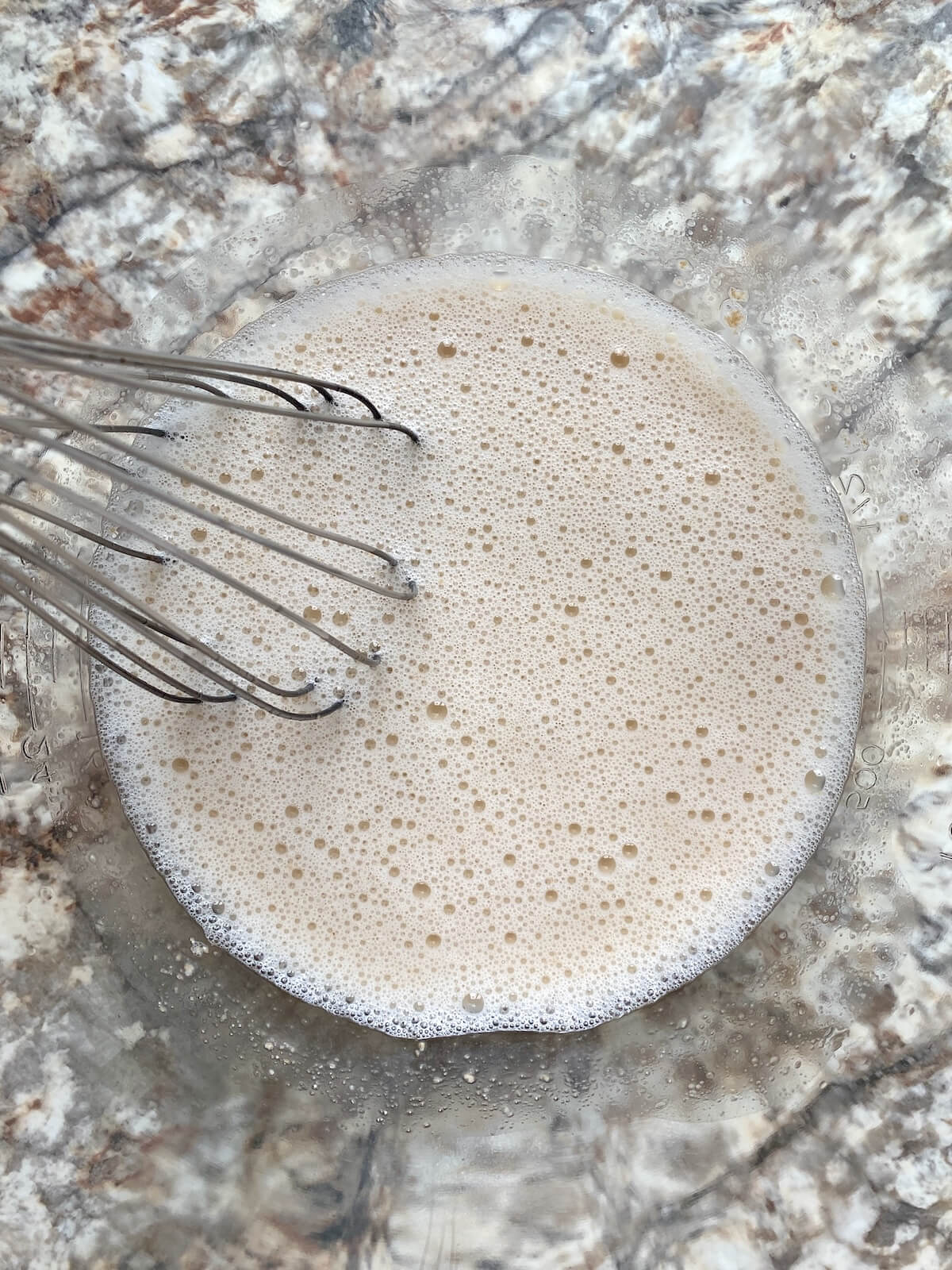 Water and active sourdough starter mixed together in a glass mixing bowl with a wire whisk sticking out to the left.