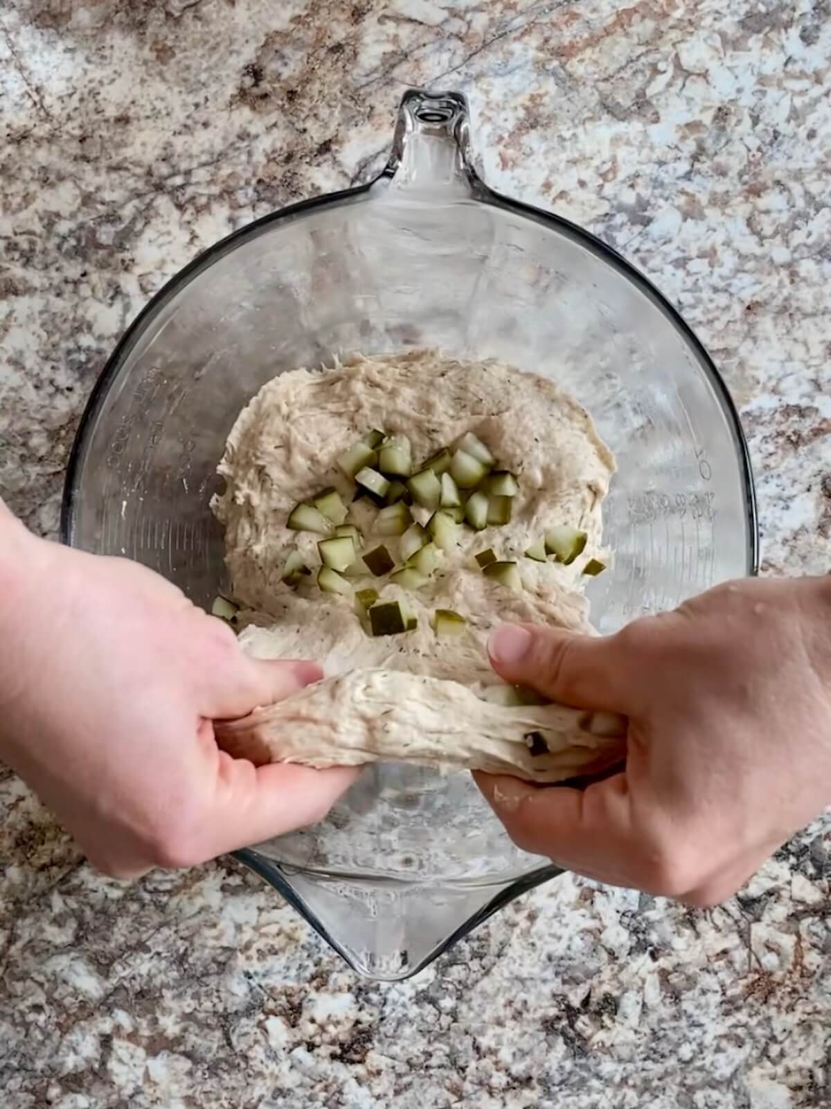 Two hands stretching and folding the chopped pickles into the dough.