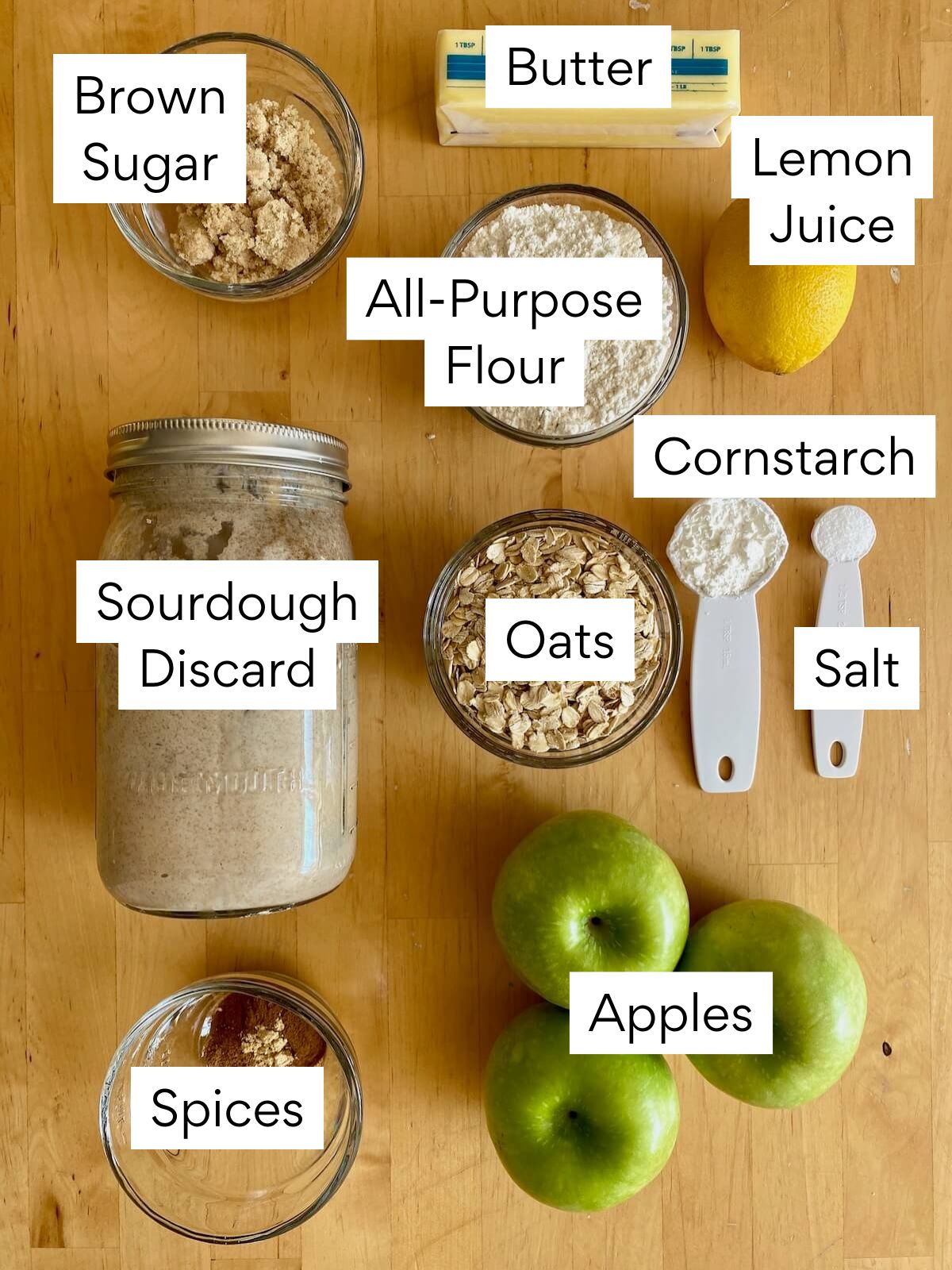 The ingredients to make sourdough apple crisp. Each ingredient is labeled with text. They include brown sugar, butter, all-purpose flour, lemon juice, sourdough discard, oats, cornstarch, salt, spices, apples.