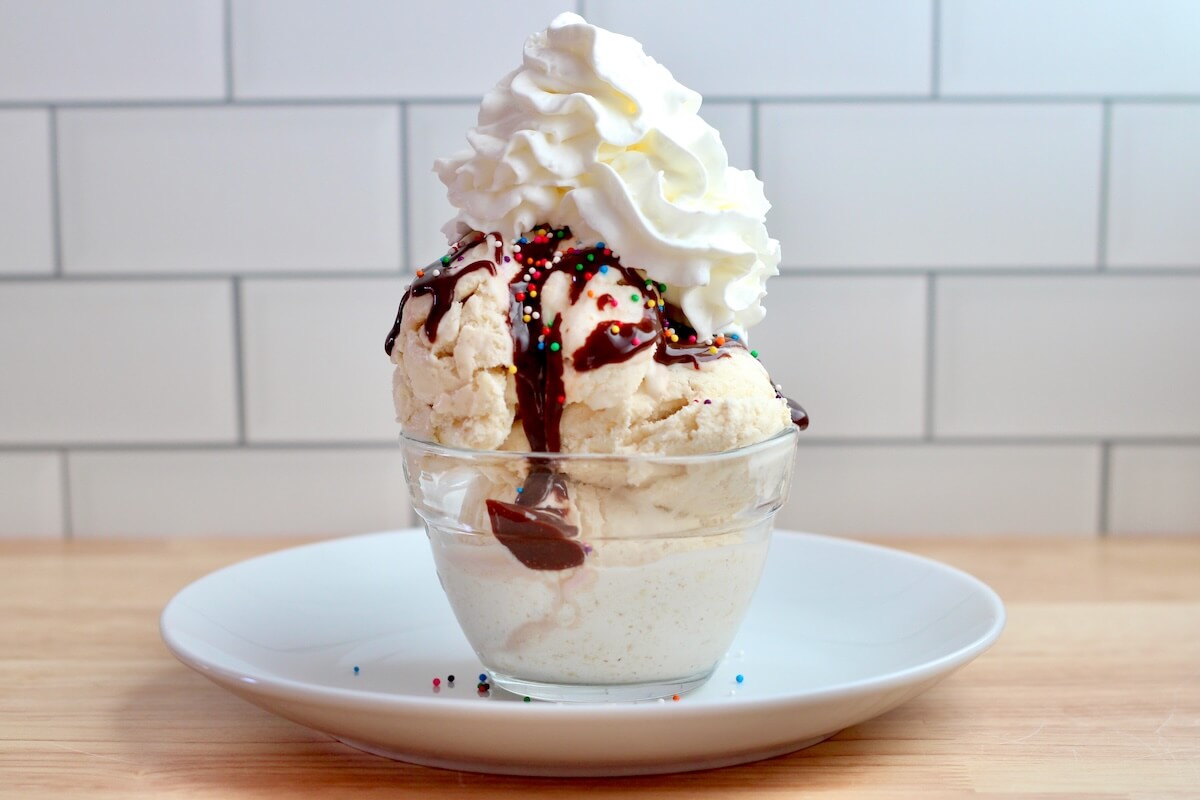 A sourdough ice cream sundae in a small bowl sitting on top of a plate. The sundae has ice cream, hot fudge, whipped cream, and colorful sprinkles.