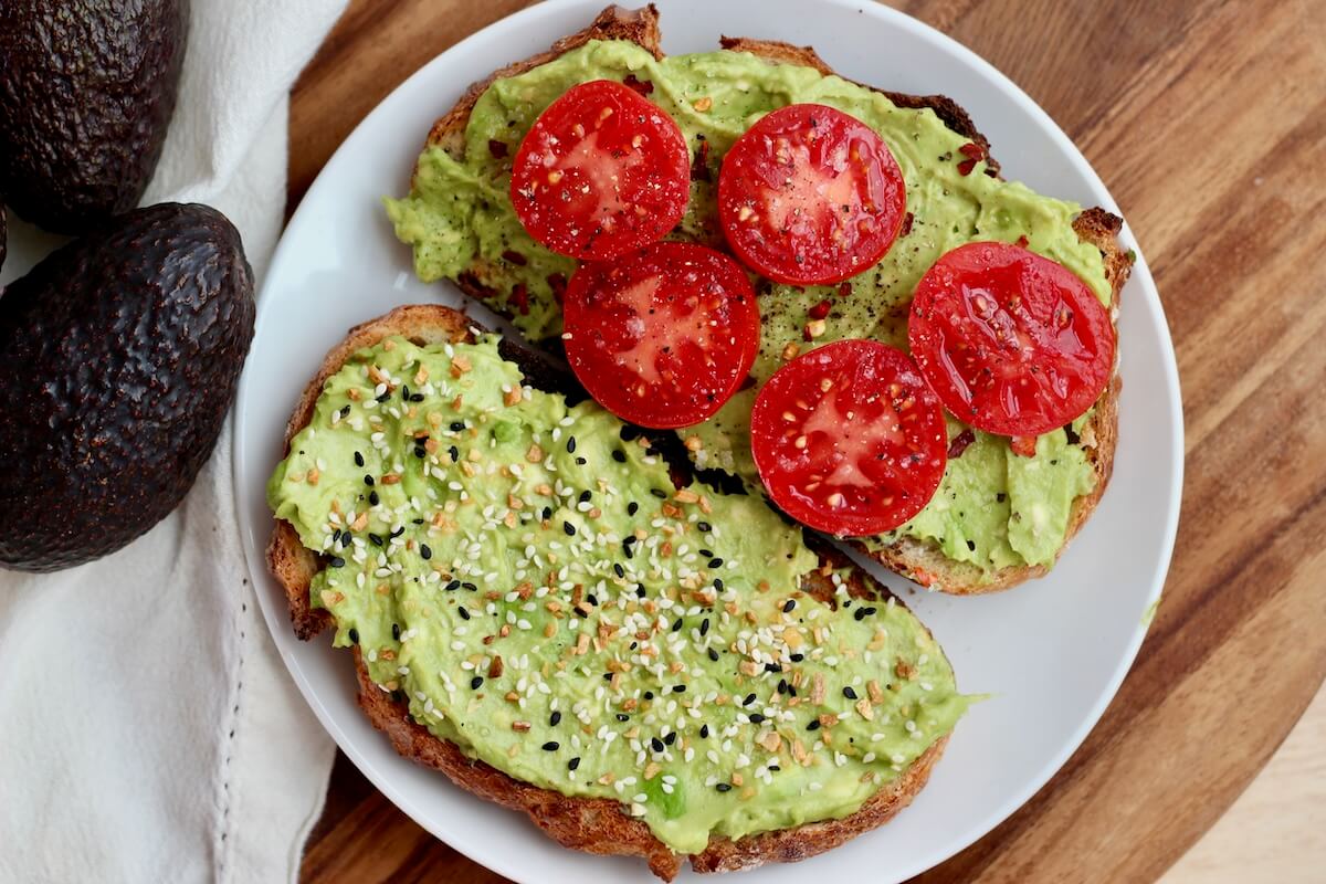 Two pieces of sourdough avocado toast on a small plate. One is topped with everything bagel seasoning and the other is topped with fresh tomato slices and seasonings.