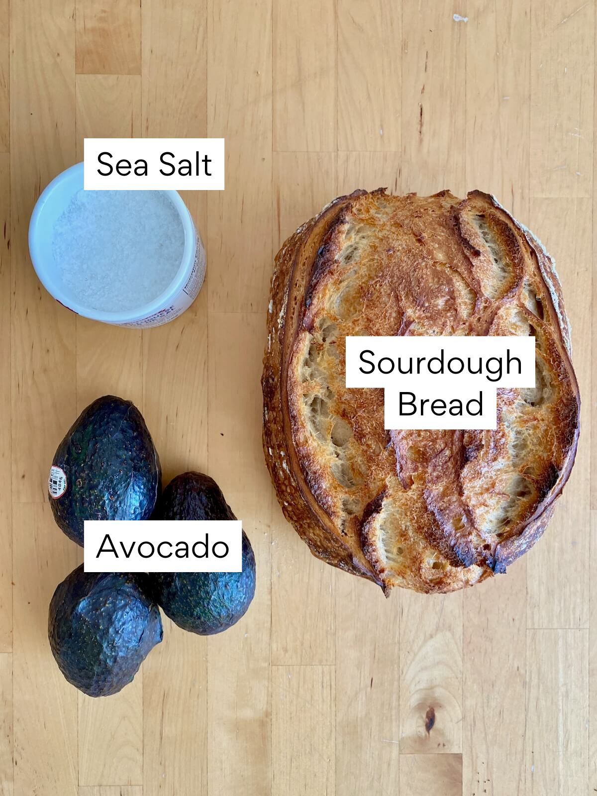 The ingredients to make sourdough avocado toast. Each ingredient is labeled with text. They include sourdough bread, avocado, and sea salt.