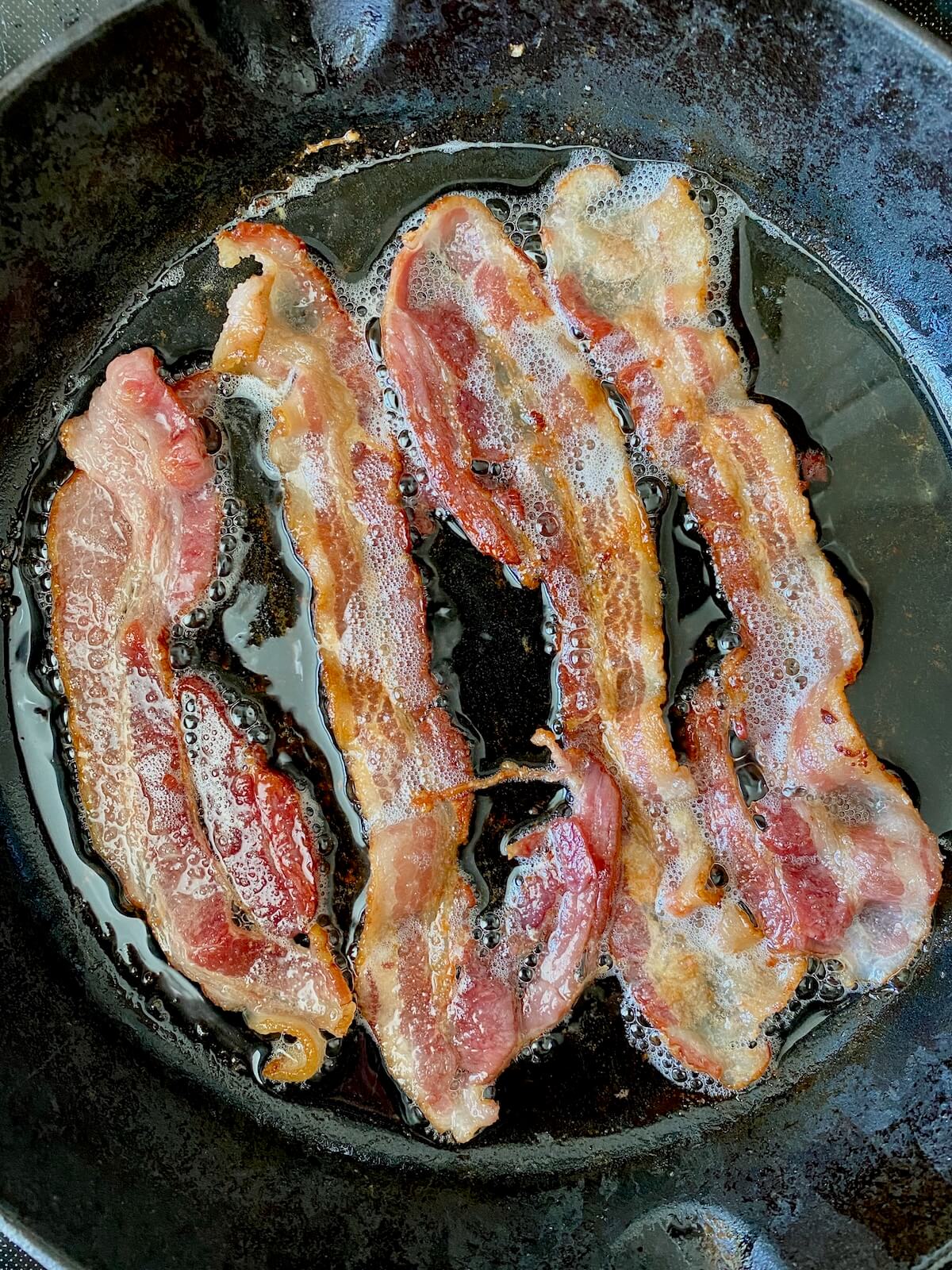 Four slices of bacon cooking in a cast iron skillet.