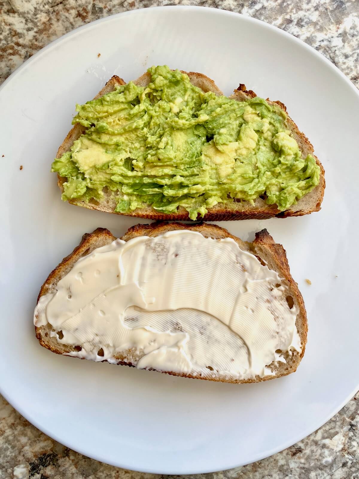 Two slices of sourdough bread on a small white plate. One piece has mashed avocado on it and the other piece has mayonnaise on it.