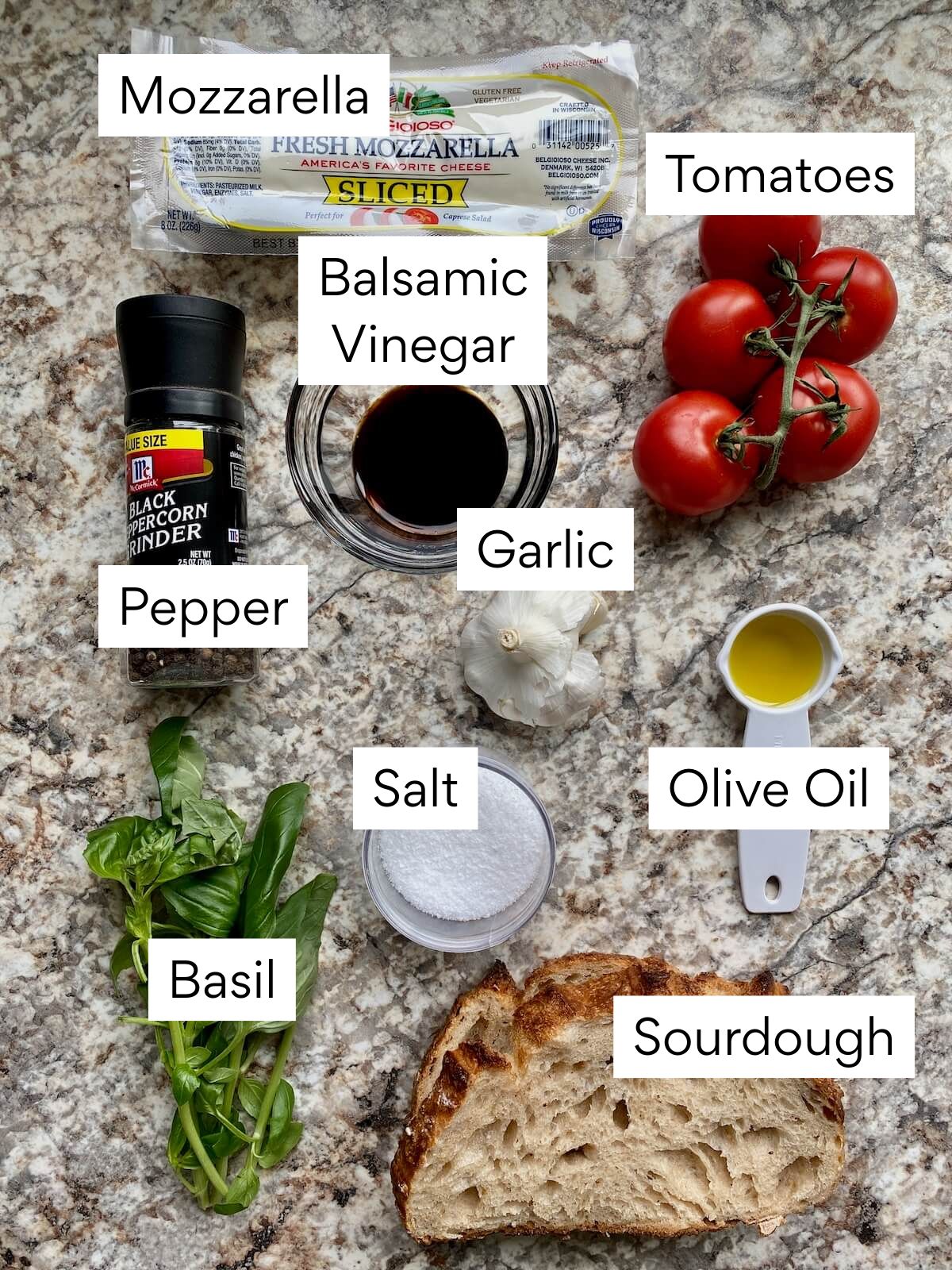 The ingredients to make sourdough bruschetta. Each ingredient is labeled with text. They include mozzarella, tomatoes, balsamic vinegar, garlic, pepper, salt, olive oil, basil, and sourdough bread.