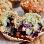 A sourdough blueberry muffin split in half on a muffin liner. There are several more whole muffins out of focus in the background.