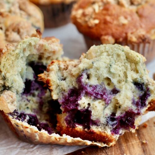 A sourdough blueberry muffin split in half on a muffin liner. There are several more whole muffins out of focus in the background.