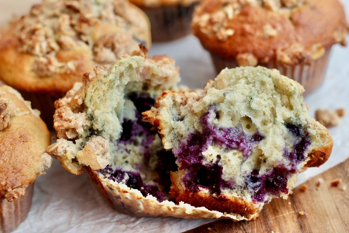 A sourdough blueberry muffin split in half on a muffin liner. There are several more whole muffins out of focus in the background.