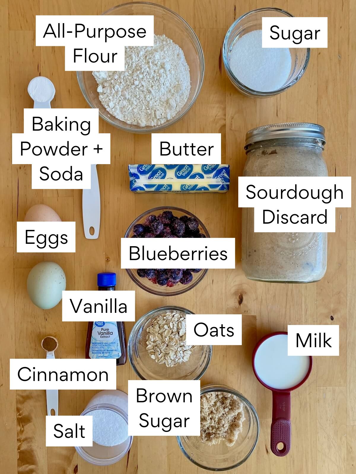 The ingredients to make sourdough blueberry muffins. Each ingredient is labeled with text. They include all-purpose flour, sugar, butter, baking powder, baking soda, sourdough discard, eggs, blueberries, vanilla, oats, cinnamon, brown sugar, milk, salt.