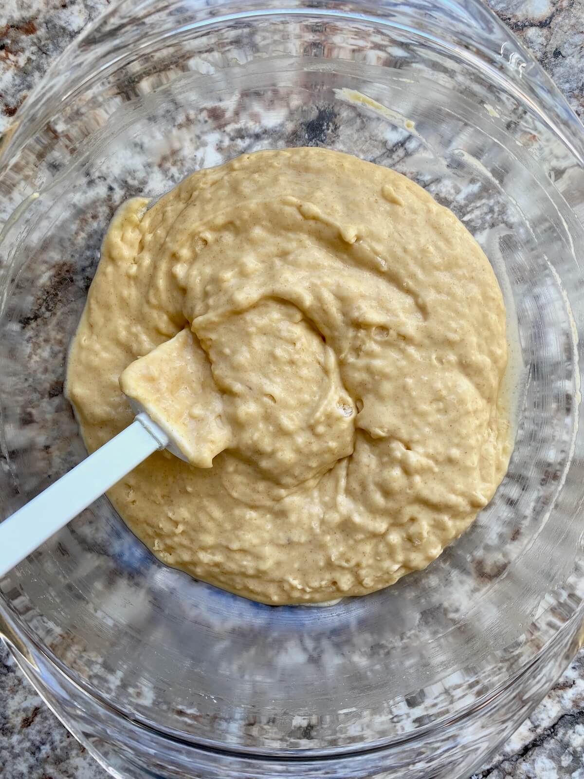 The muffin batter mixed together in a glass bowl with a rubber spatula sticking out to the left.