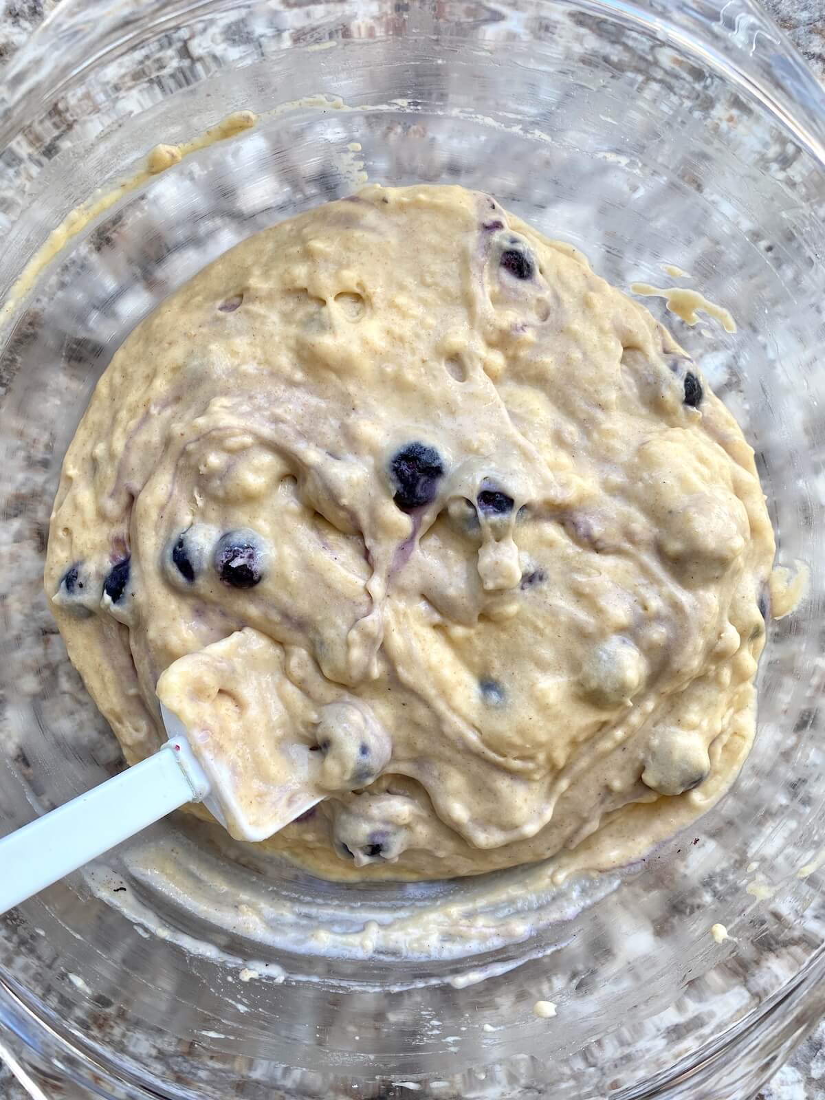 The muffin batter with frozen blueberries mixed into it in a glass bowl with a rubber spatula sticking out to the left.
