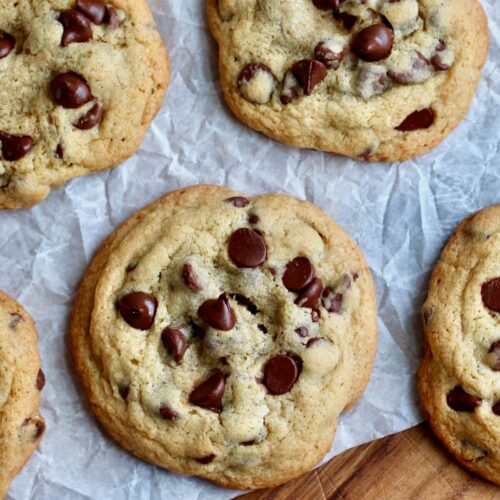 Several sourdough chocolate chip cookies on a piece of wrinkled parchment paper on a wooden cutting board.